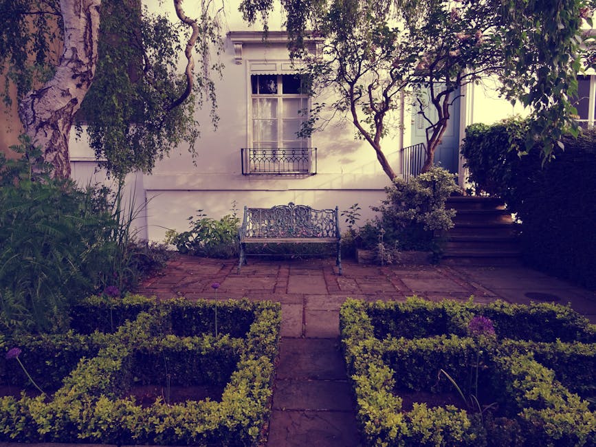 A small, well-maintained garden in front of a white house, featuring a paved stone patio with a decorative metal garden bench positioned centrally against the house wall. Surrounding the patio are nea