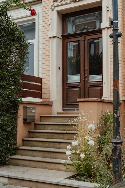 The image shows the front entrance of a traditional brick townhouse with a set of beige stone steps leading up to a wooden front door featuring glass panels. To the left of the steps, there is a lush,
